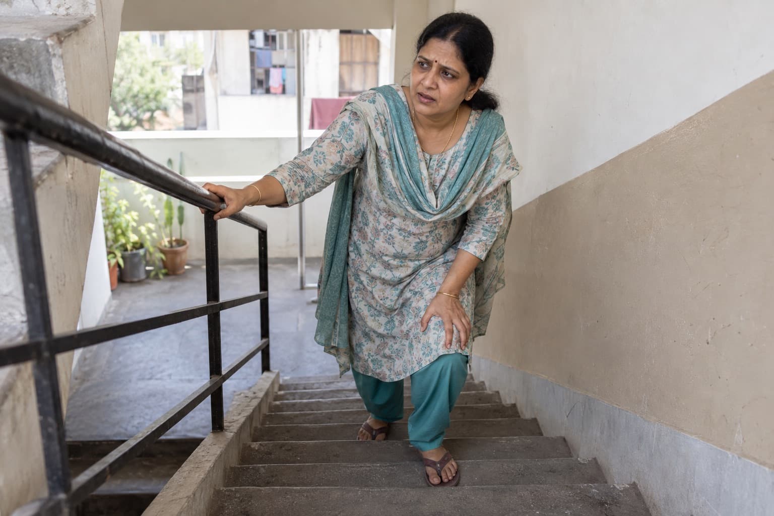 An Indian woman pauses on the stairs of a modest apartment building because her knee hurts
